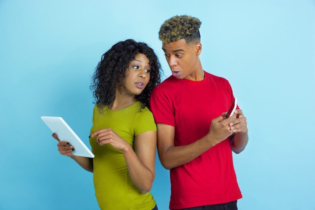 Free photo young emotional african-american man and woman in colorful clothes using gadgets, peeping on screens on blue b