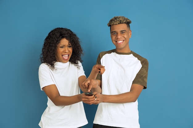 Young emotional african-american man and woman posing on blue background. Beautiful couple. Concept of human emotions, facial expession, relations, ad. Watch TV together, his favourite channel.