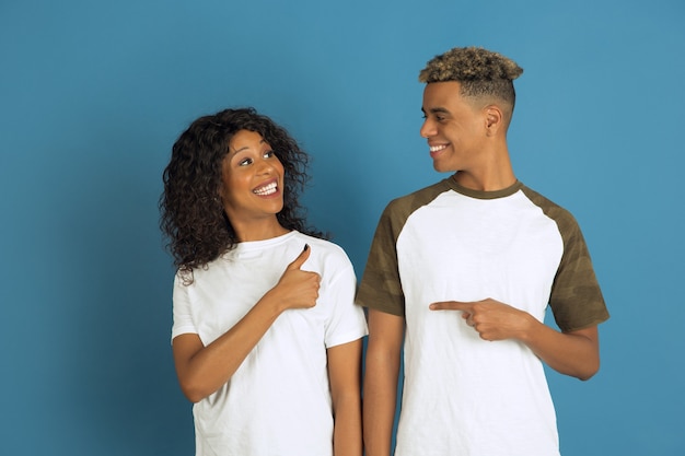 Young emotional african-american man and woman in white casual clothes posing on blue background. Beautiful couple. Concept of human emotions, facial expession, relations, ad. Pointing on each other.