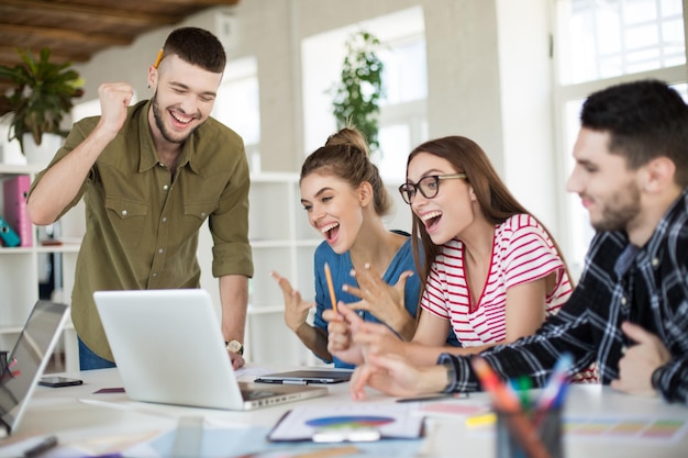 young emotional business people joyfully working on laptop together group of laughing men and women spending time at work in modern cozy office