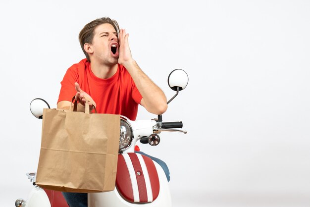 Young emotional courier guy in red uniform sitting on scooter holding paper bag calling someone on white wall
