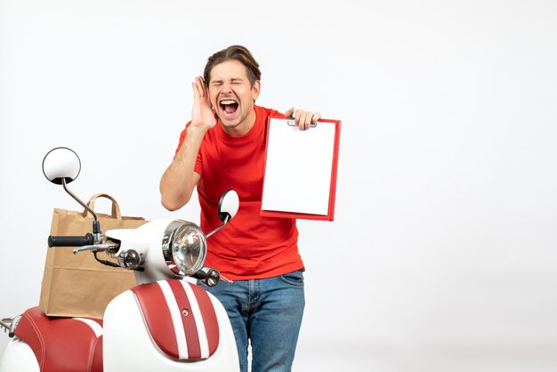 Young emotional happy smiling delivery guy in red uniform standing near scooter showing document on white wall