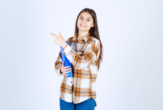 Young employee with blue folder pointing at her side on white wall. 