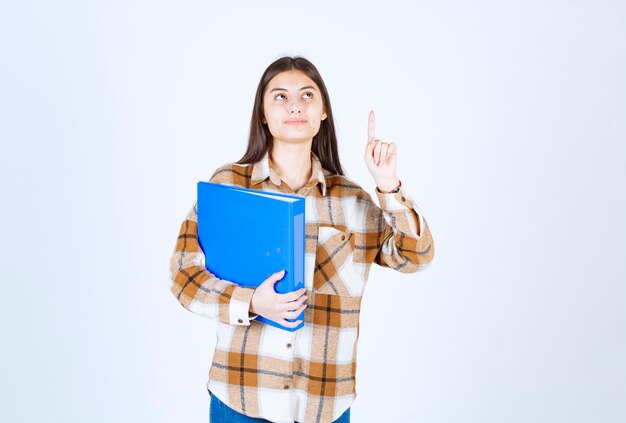 Young employee with blue folder pointing at upside on white wall. 