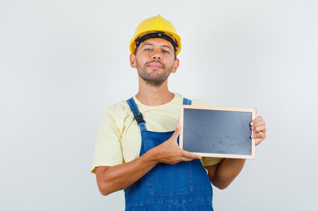 Giovane assistente tecnico che tiene lavagna e sorridente in uniforme, vista frontale.