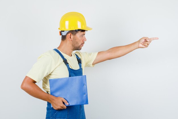 Young engineer holding clipboard and pointing to side in uniform front view.