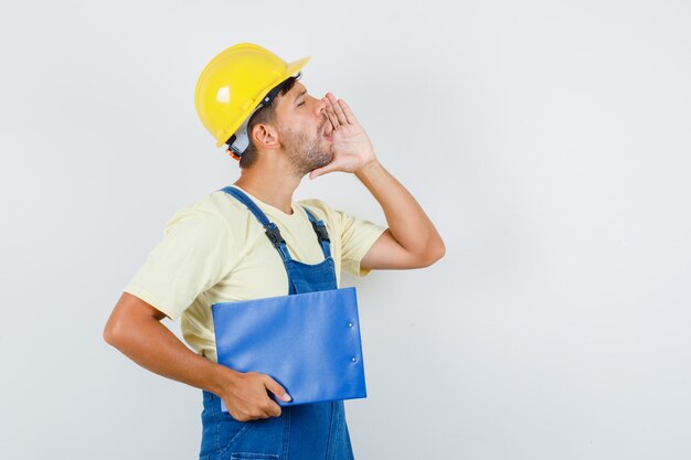 Young engineer holding clipboard and shouting in uniform front view.