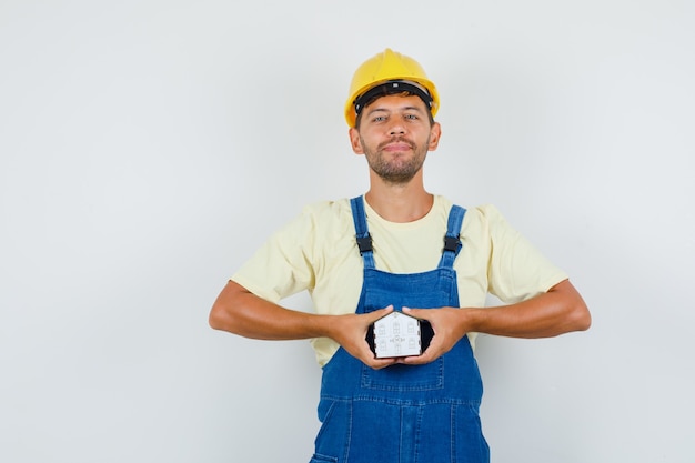 Foto gratuita giovane ingegnere che tiene il modello di casa in uniforme e sembra allegro, vista frontale.