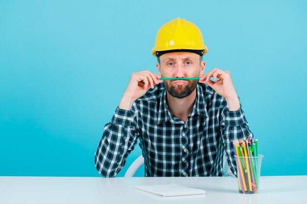 Young engineer man is holding pencil between nose and lips on blue background