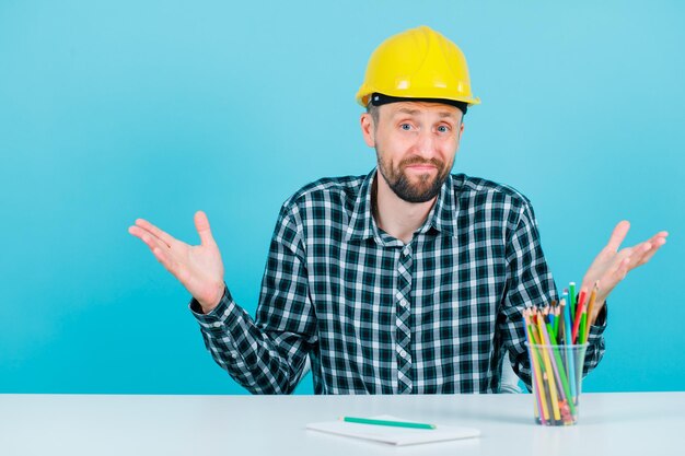 Young engineer man is looking at camera by opening wide his hands on blue background