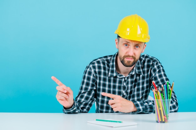 Young engineer man is pointing left with forefingers on blue background