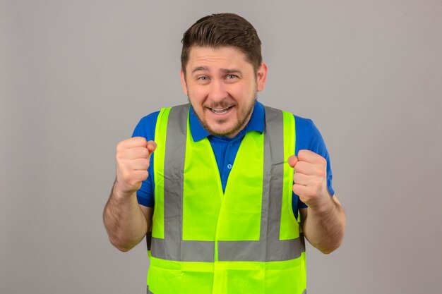 Young engineer man wearing construction vest clenching fists smiling standing with happy face over isolated white background