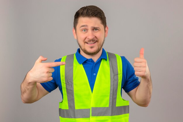 Young engineer man wearing construction vest looking at camera smiling cheerfuly showing thumb up pointing with finger standing over isolated white background