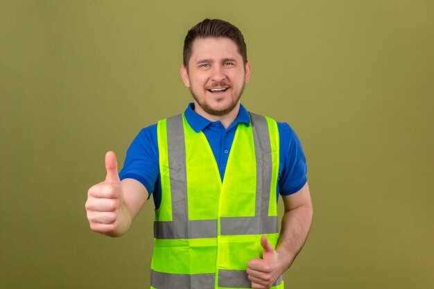 Young engineer man wearing construction vest looking at camera with big smile and happy face showing thumb up over isolated green background