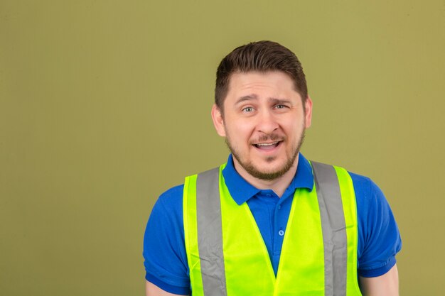Young engineer man wearing construction vest looking confused with expression as asking question over isolated green background