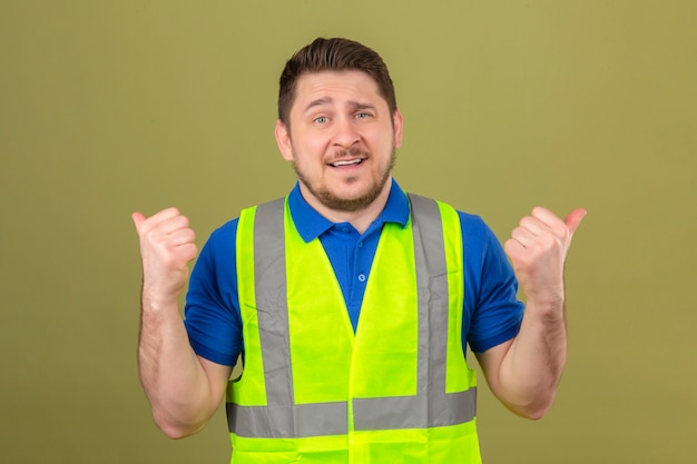 Young engineer man wearing construction vest pointing to the back behind with hands and thumbs up smiling over isolated green background