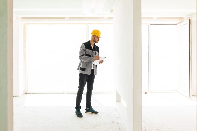 Young engineer in work clothes and yellow hardhat holding plan of new apartments thoughtfully looking on wall with big window on background