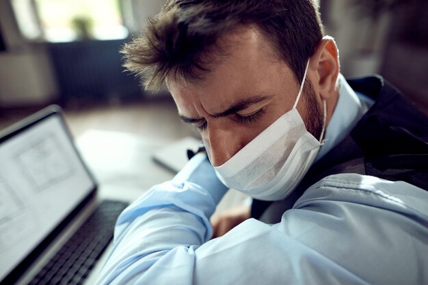 Young entrepreneur coughing into elbow while wearing protective face mask in the office