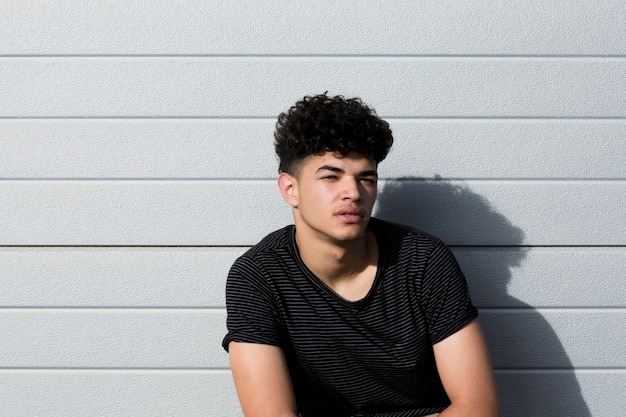 Young ethnic curly man in black shirt sitting against grey wall