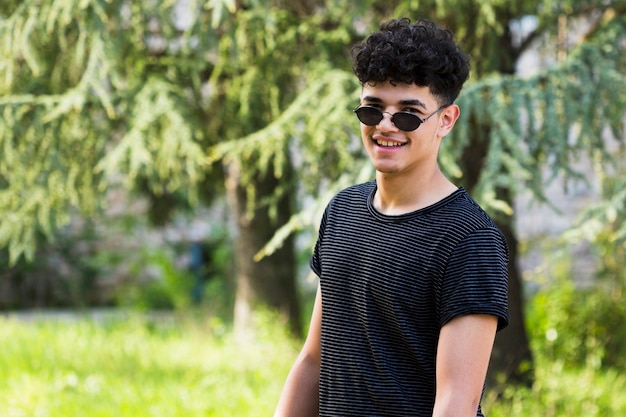 Foto gratuita giovane uomo riccio etnico in camicia nera e occhiali da sole su sfondo di alberi