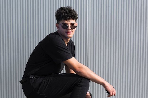 Young ethnic curly man in sunglasses and black clothes against grey wall