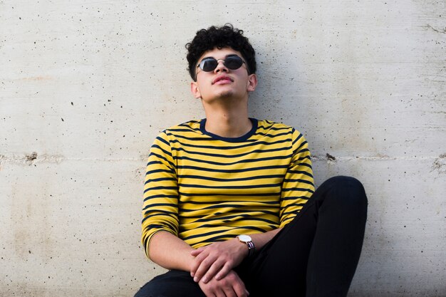 Young ethnic curly man in sunglasses leaning on grey concrete wall