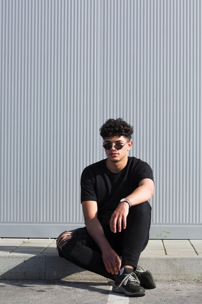 Young ethnic man in sunglasses sitting against grey wall