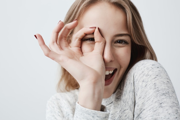 OKジェスチャーで指を通して見る若いヨーロッパのうれしそうな金髪の女性。広く笑っているカジュアルな服装の女性。彼女の幸せな顔の表情は、すべてが計画通りに進んでいることを証明しています。