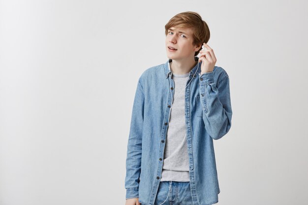 Young european man with fair hair in denim shirt, listens to music on mobile phones, wearing white earphones. Young male enjoys favourite songs, uses wifi. Modern techologies concept