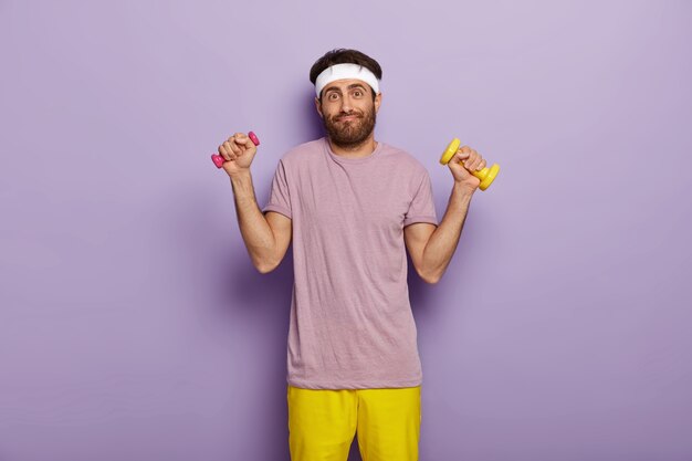 Young European man works on muscles, raises arms with dumbbells, has exercises indoor, has athletic body, dressed in purple t shirt and yellow shorts, stands indoor, has funny look, dark bristle