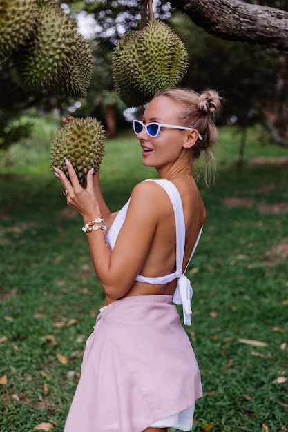 Бесплатное фото Молодой европейский довольно стильный модный блоггер туристическая женщина с фруктами дуриан джек у дерева на поле