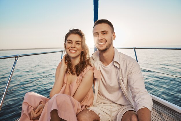 Young european romantic couple smiling while sitting at bow of boat, hugging, enjoying their holidays. Two close friends recently became something more to each other