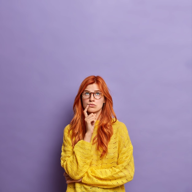 young European woman in glasses with natural red hair stands in thoughtful pose tries to choose something or thinks about future concentrated above.