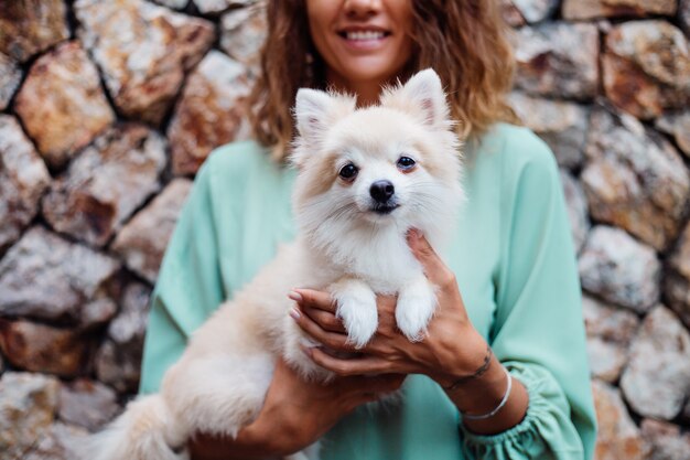 Young european woman in romantic summer dress bracelet holds cute fluffy pomeranian puppy spitz outside villa