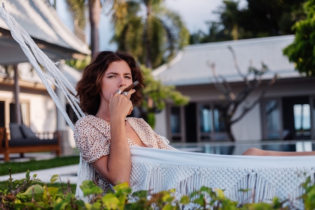 熱帯の高級ヴィラホテル、夕日の自然光の外のハンモックに横たわって葉巻を吸う若いヨーロッパの女性