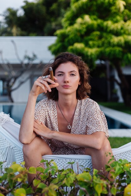 Молодая европейская женщина курит сигару, лежа на гамаке возле роскошного тропического отеля на вилле, закат естественный свет