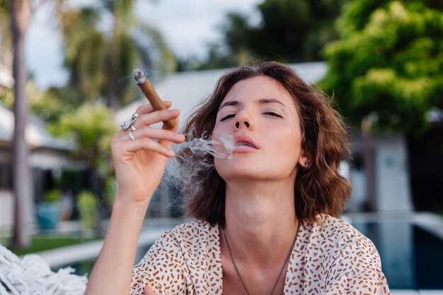 열대 럭셔리 빌라 호텔, 일몰 자연 채광 외부 해먹에 누워 젊은 유럽 여성 흡연 시가