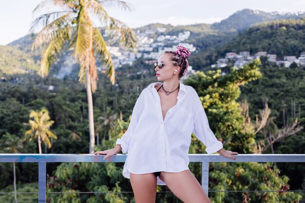 Young european woman in white long shirt black bikini with pink braids on head on tropical palm trees