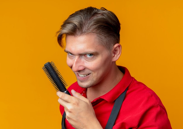 Young evil joyful blonde male barber in uniform holds hair comb isolated on orange space with copy space
