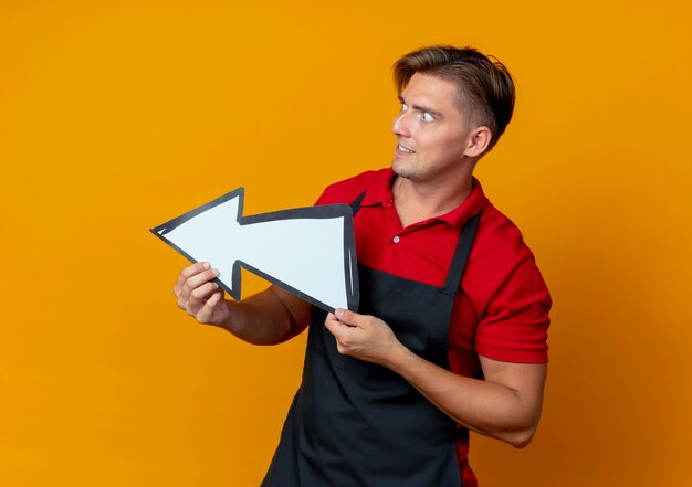 Young excited blonde male barber in uniform holds direction mark and looks at side isolated on orange space with copy space