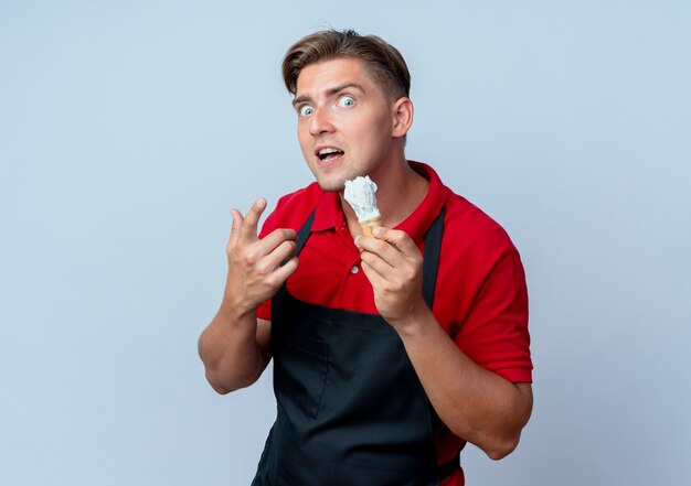 young excited blonde male barber in uniform holds shaving brush isolated on white background