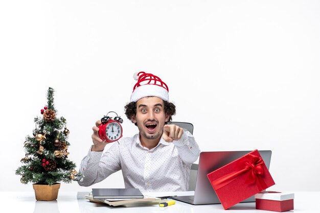 Young excited business person with santa claus hat and showing clock working alone sitting in the office on white background