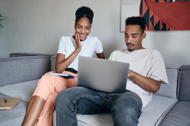 Young excited casual African American couple happily working on laptop together on sofa at modern home