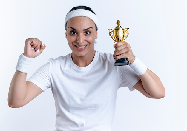 Young excited caucasian sporty woman wearing headband and wristbands holds winner cup and raises fist isolated on white space with copy space