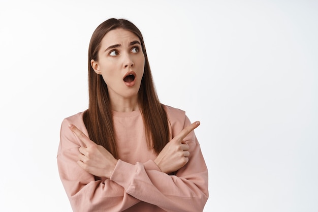 Young excited girl, gasping amazed, pointing sideways two ways choices makes choice, standing on white