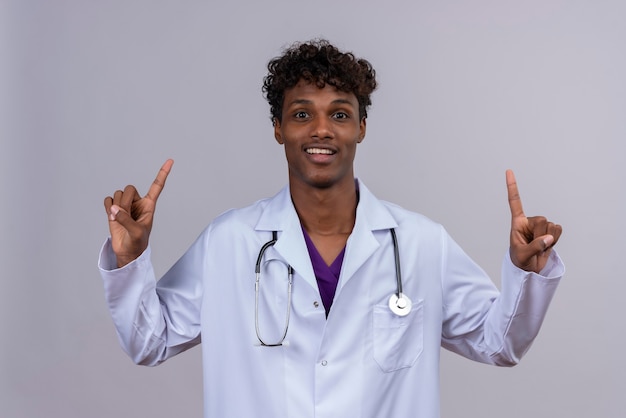 A young excited handsome dark-skinned male doctor with curly hair wearing white coat with stethoscope pointing up with index fingers 