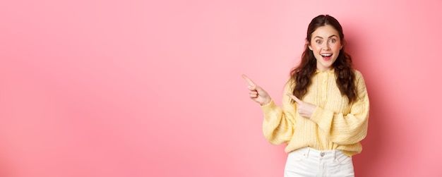 Free photo young excited woman in casual outfit showing banner pointing fingers left aside copy space standing against pink background