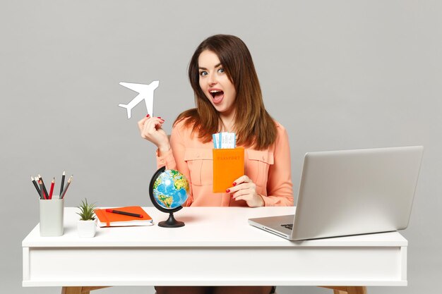 종이 비행기, 지구 세계 지구, 여권 탑승권 티켓을 들고 있는 젊은 흥분한 여성이 회색 배경에 격리된 pc 노트북과 함께 책상에서 일합니다. 성취 비즈니스 경력 라이프 스타일 개념입니다.