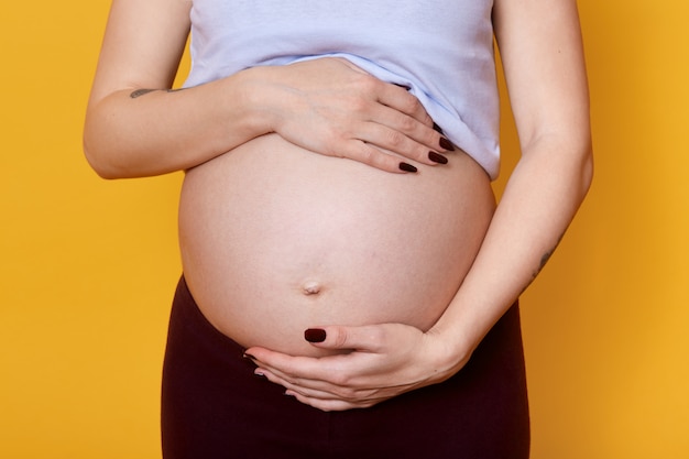 若い顔のない妊娠中の女性は彼女の大きなお腹を手で保持しています。写真で撮影されている妊娠中のモデル