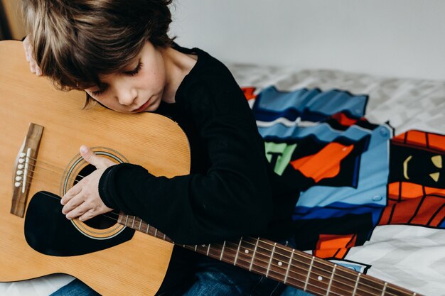 Young fair-haired boy sitting on the bed and holding a guitar